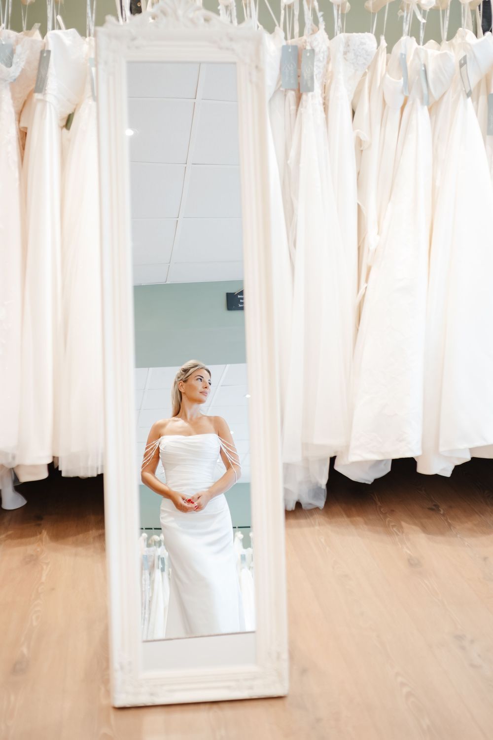Bride tries on white strapless wedding gown in bridal boutique, reflected in ornate floor mirror surrounded by dresses.