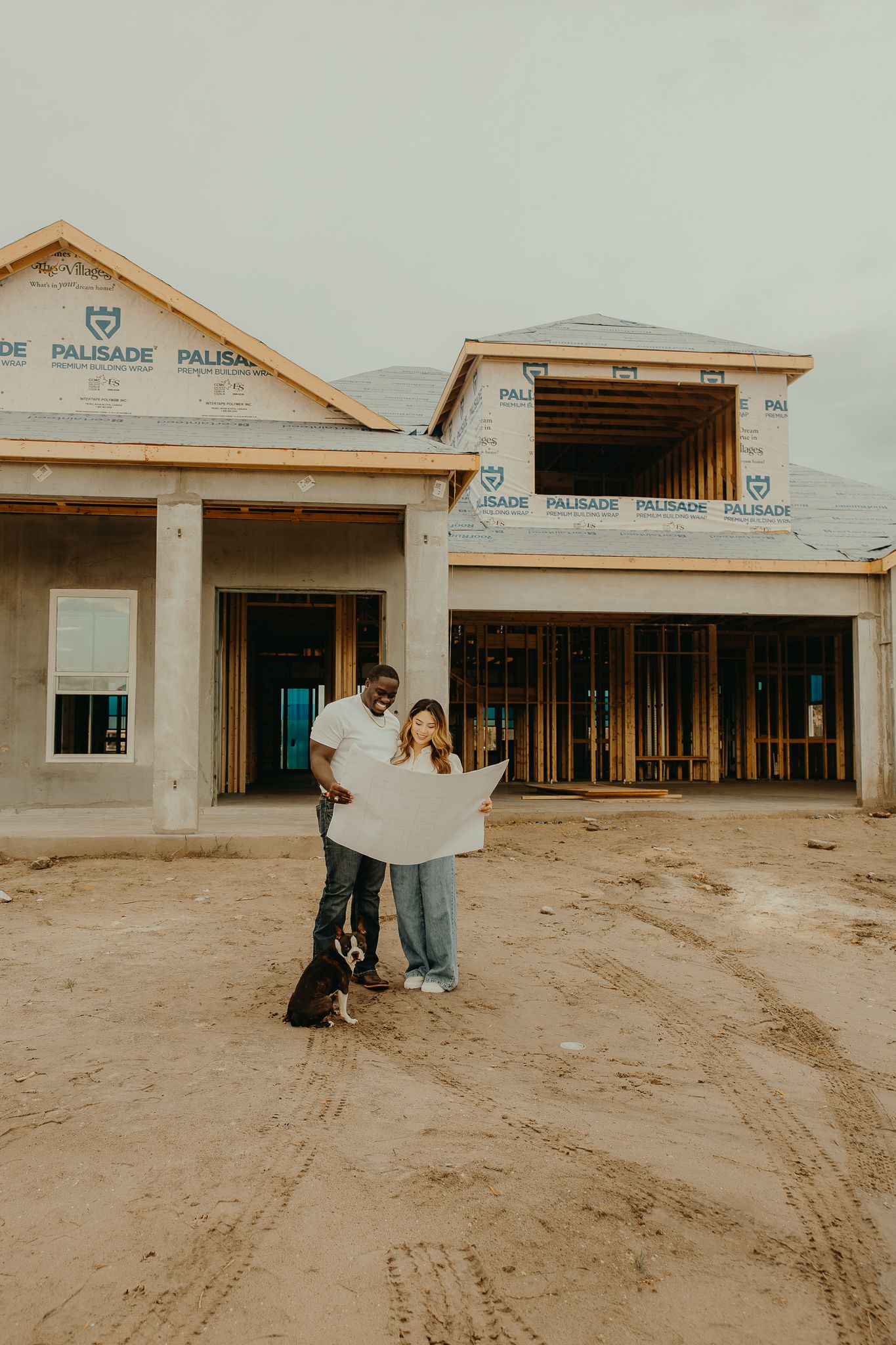 Couple holding blueprints with dog in front of new home under construction with Palisade wrap visible.