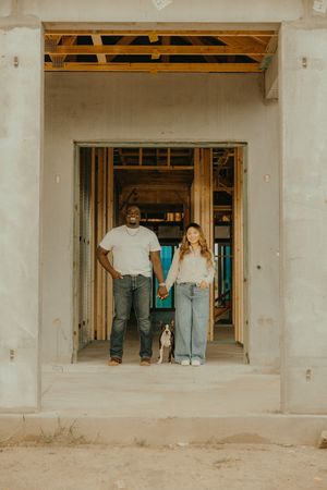 Couple holding hands with their dog in the doorway of a home under construction.