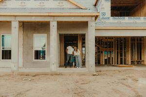 Couple standing in doorway of home under construction with exposed wooden framing and sandy ground.