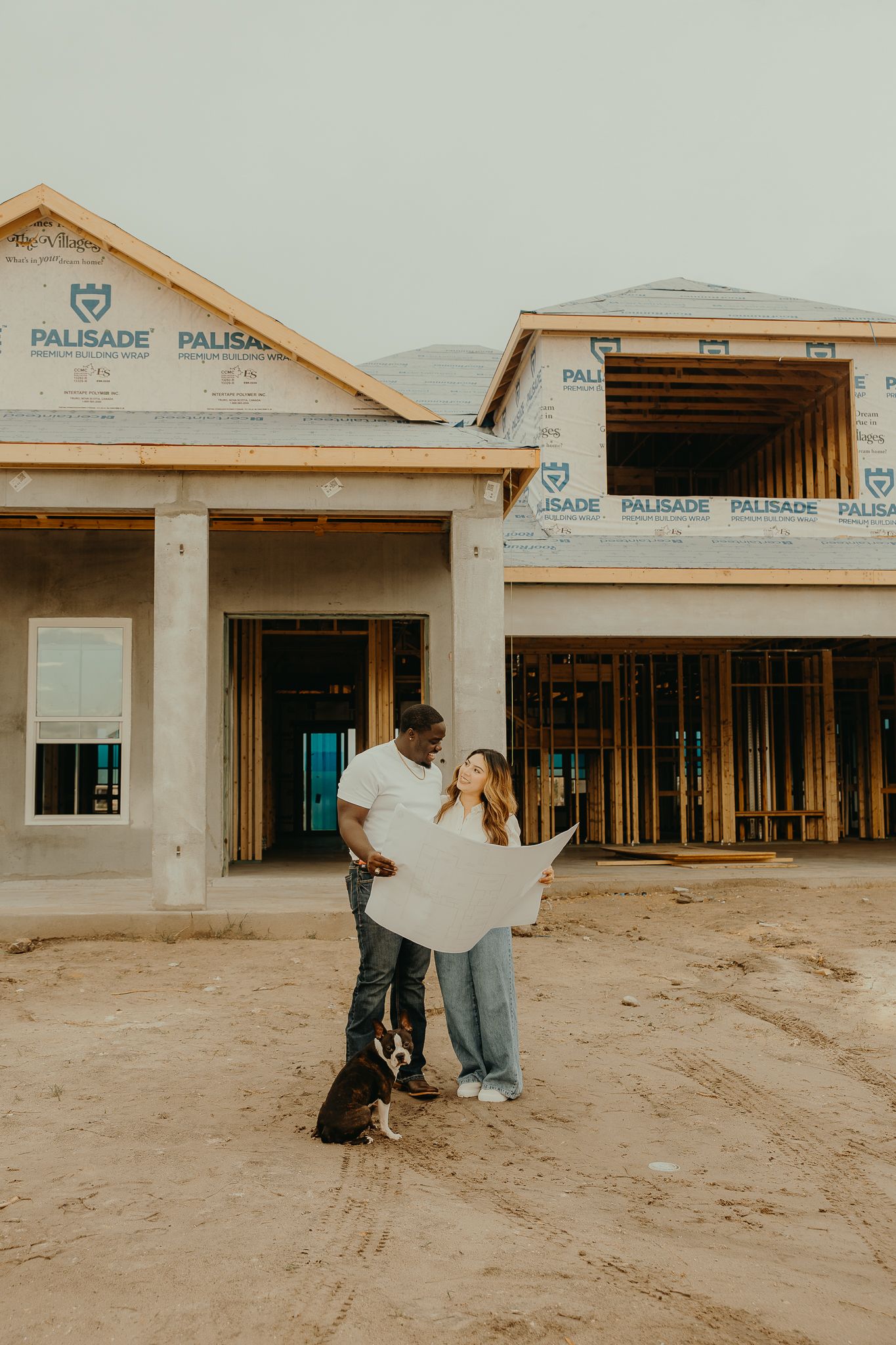 Couple holding blueprints with dog in front of new home under construction with Palisade wrap visible.