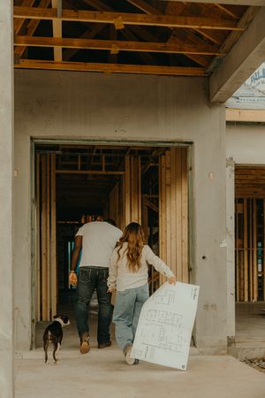 Couple with dog reviewing blueprints at a home construction site with wood framing and concrete walls.