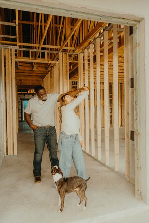 Couple dancing and laughing with their dog inside a new home under construction with wooden framing.