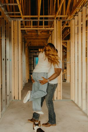 Couple embracing and lifting each other joyfully inside a new home under construction with wooden framing.