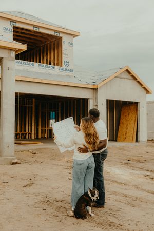Couple reviewing blueprints with dog at new home construction site, framed house in background.