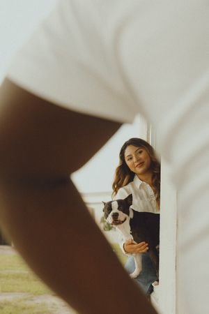 Woman smiling and holding a Boston Terrier dog, viewed through a gap between two white walls in warm sunlight.