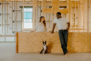 Interracial couple with Boston Terrier dog posing inside unfinished new construction home with wood framing.