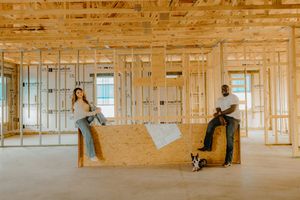 Couple sitting on plywood in a new home under construction with wood framing and a small dog nearby.