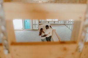 Couple embracing in an unfinished home interior, viewed through wooden framing during a new construction house tour.