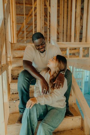 Happy interracial couple sitting on stairs inside unfinished new home, smiling and embracing warmly.