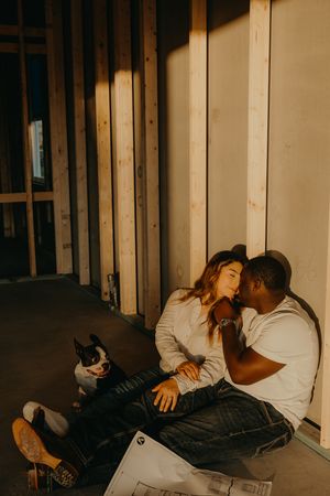 Interracial couple sitting on floor of new construction home with Boston Terrier and house blueprints.