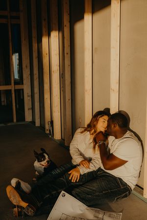 Interracial couple sitting on floor of home under construction with Boston Terrier dog in warm golden light.