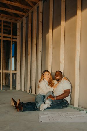 Interracial couple sitting together on floor of new construction home with blueprints, smiling at camera.
