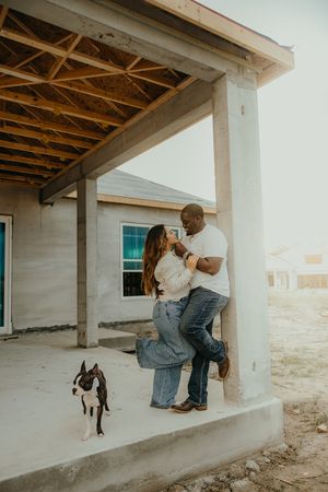 Couple embraces on the porch of their home under construction with their Boston Terrier nearby.