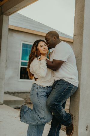 Interracial couple embracing romantically against a concrete pillar at a home construction site.