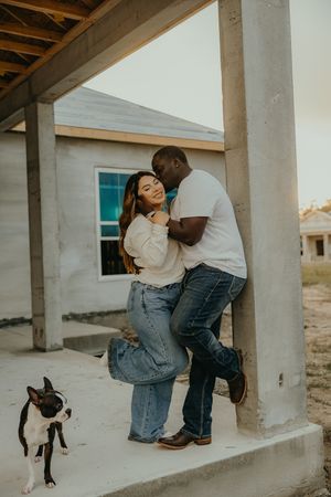 Couple embracing against a concrete pillar at a construction site with a Boston Terrier dog nearby.