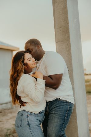 An interracial couple shares a tender embrace near a concrete pillar, both wearing white tops and jeans at golden hour.
