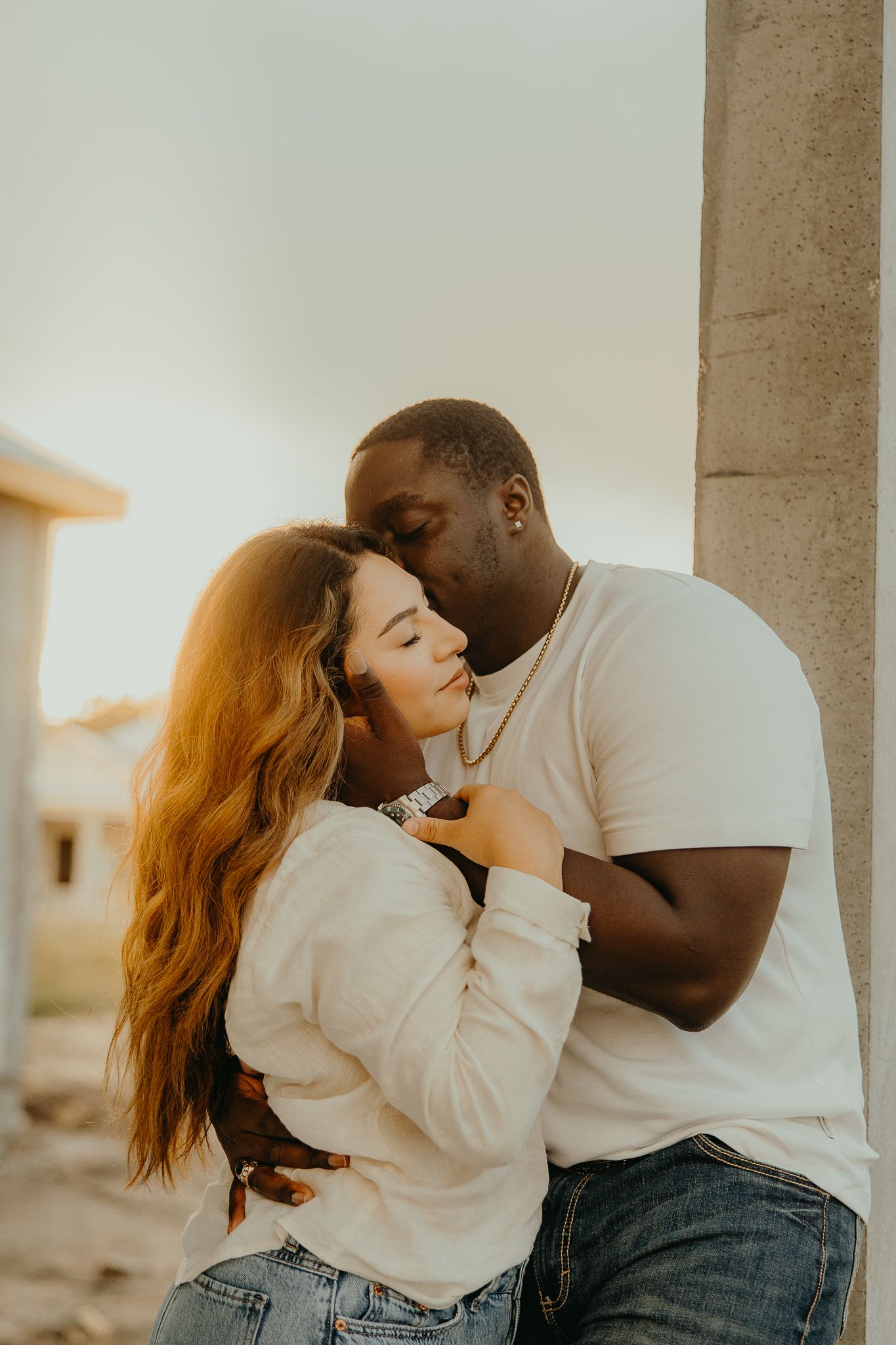 Interracial couple sharing a tender kiss while leaning against pillar of new construction home at sunset.