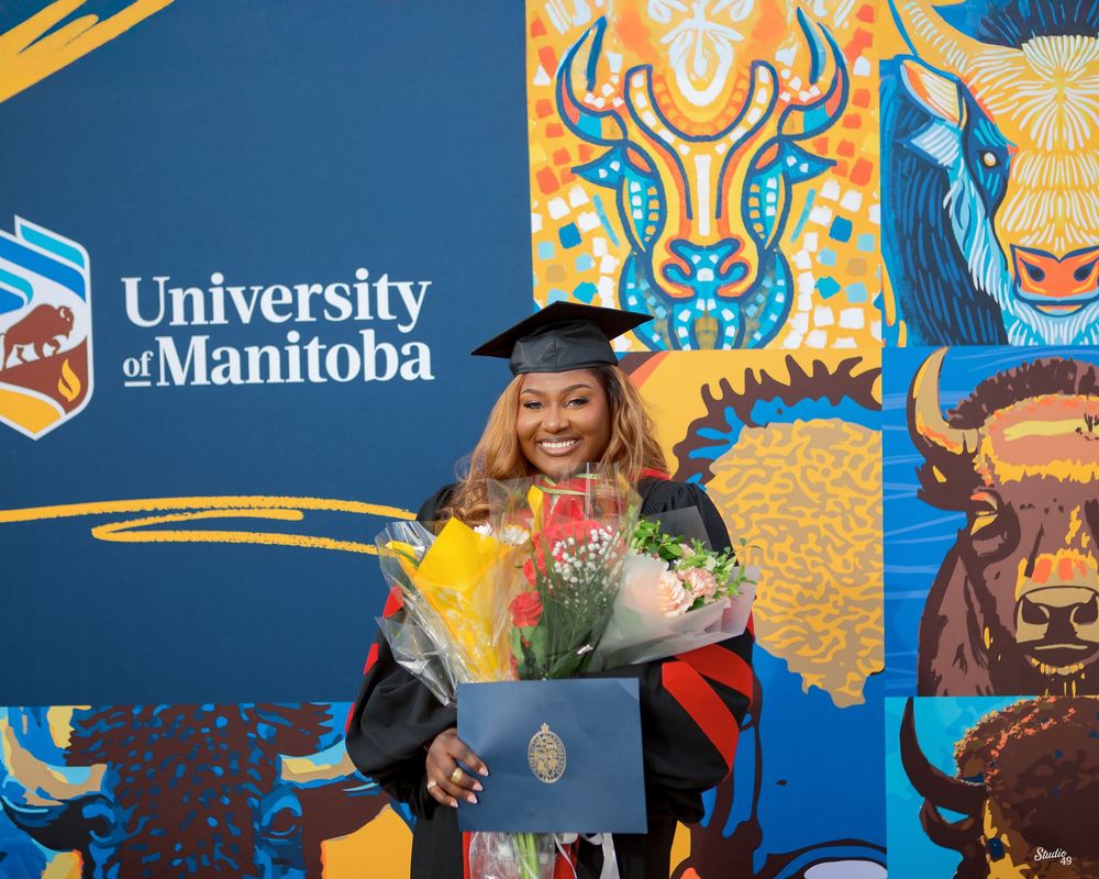 A smiling Black woman in graduation cap and gown holds flowers and diploma in front of University of Manitoba backdrop.
