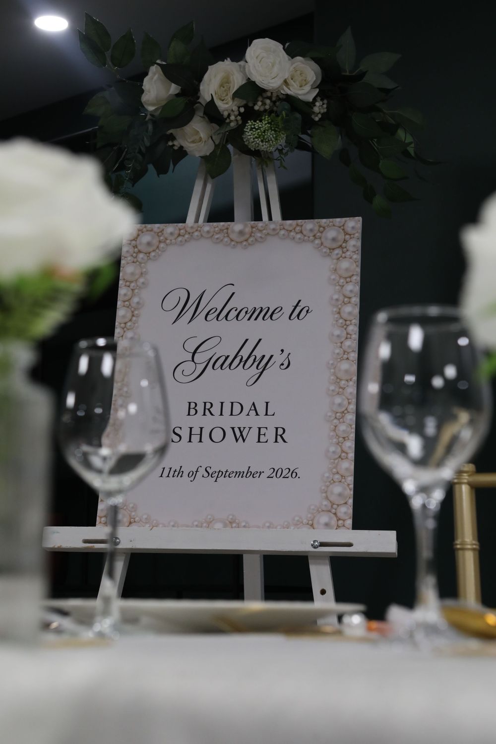 An elegant bridal shower welcome sign for Gabby on a white easel with white roses and wine glasses on a table.