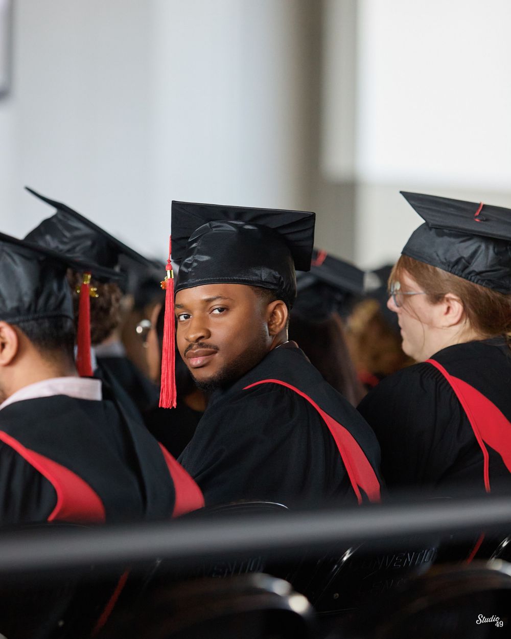 A young Black male graduate in cap and gown looks back at the camera during a university graduation ceremony.