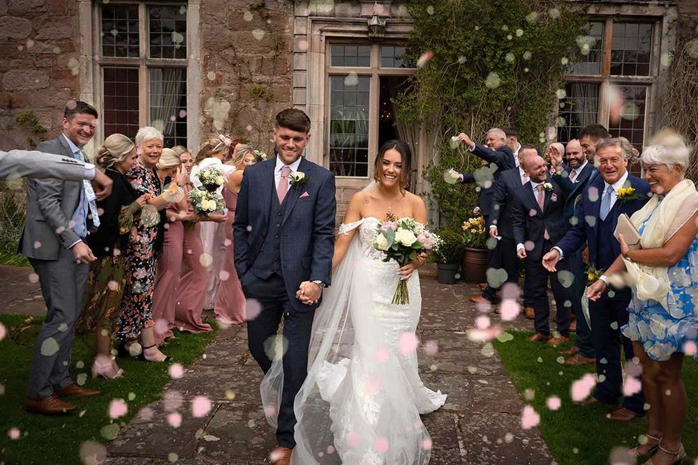 Newlywed couple walks through confetti shower outside stone venue, surrounded by cheering guests.