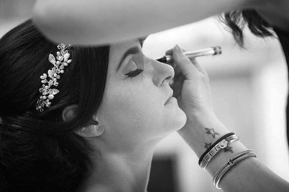 Black and white photo of a bride having makeup applied, wearing a floral hair accessory.