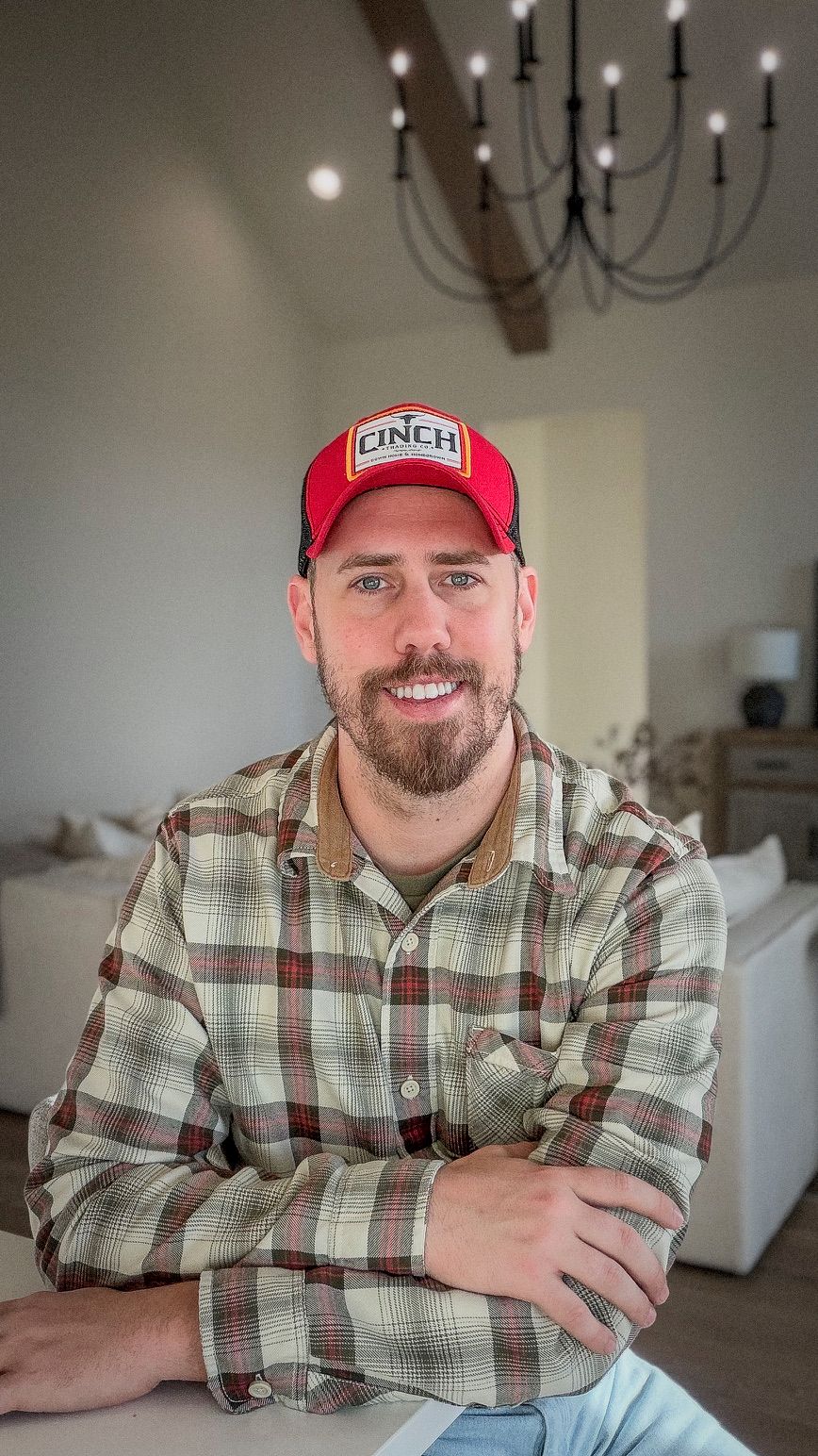 Smiling bearded man in plaid shirt and red Cinch cap in modern farmhouse interior.