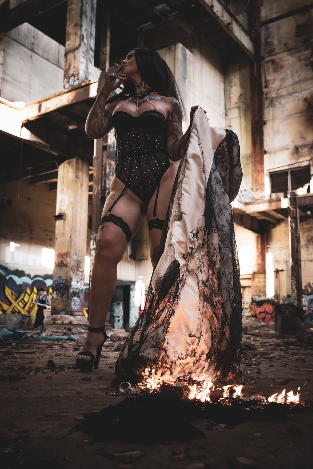 Woman in black corset lingerie poses dramatically with burning fabric in an abandoned graffiti-covered industrial building.