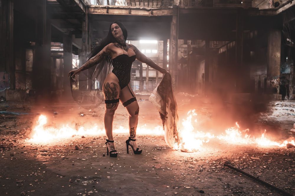Tattooed woman in black outfit poses dramatically among flames in an abandoned industrial building.
