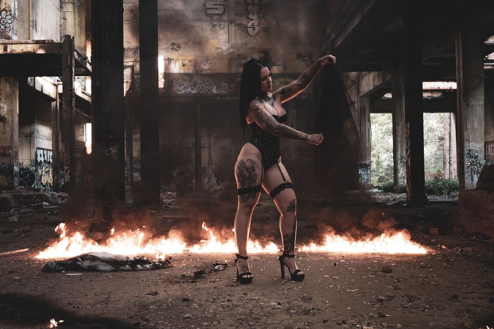A tattooed woman in dark attire poses dramatically amid flames in an abandoned graffiti-covered industrial building.