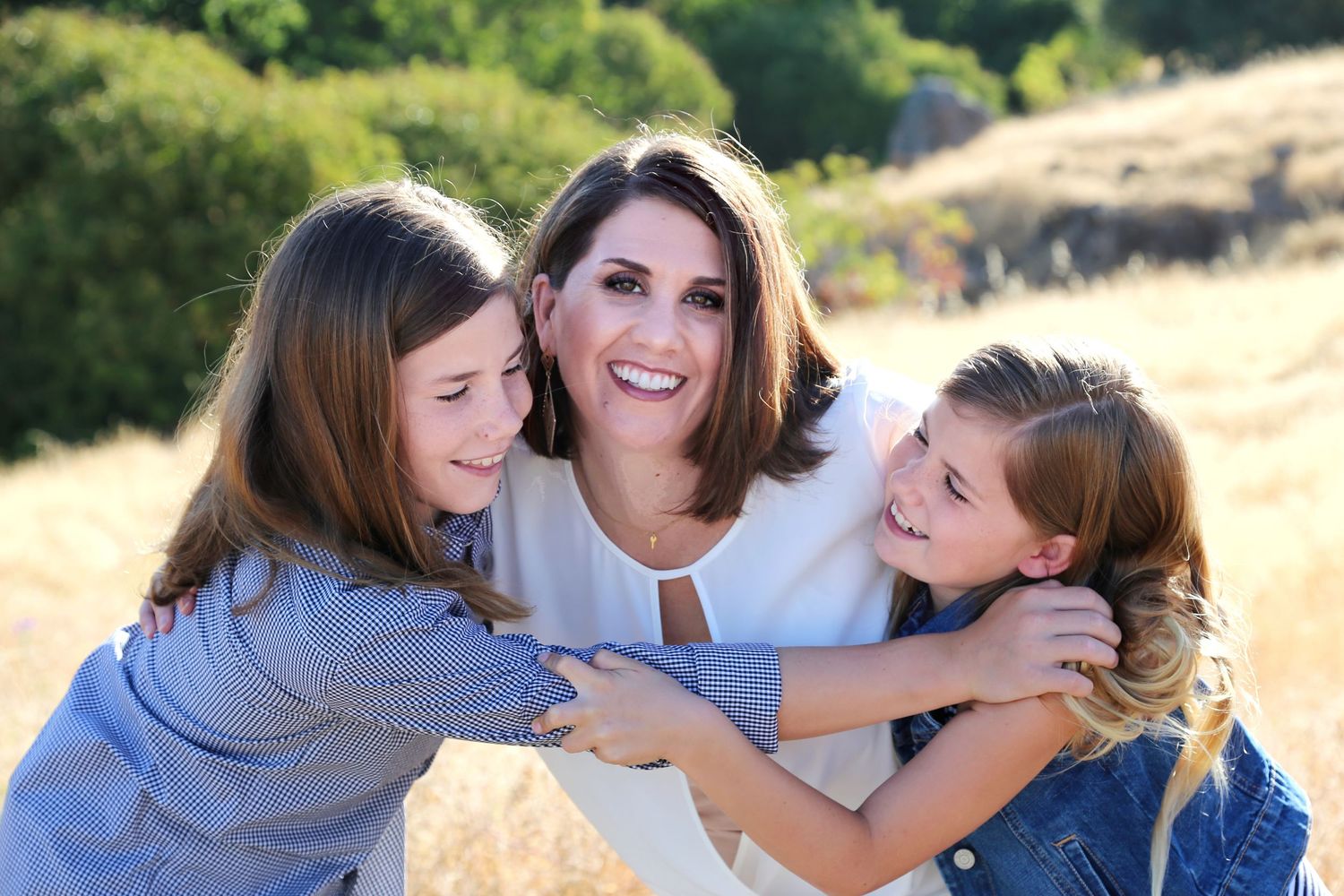 A woman smiles warmly while being hugged by two young girls in a sunny golden field.