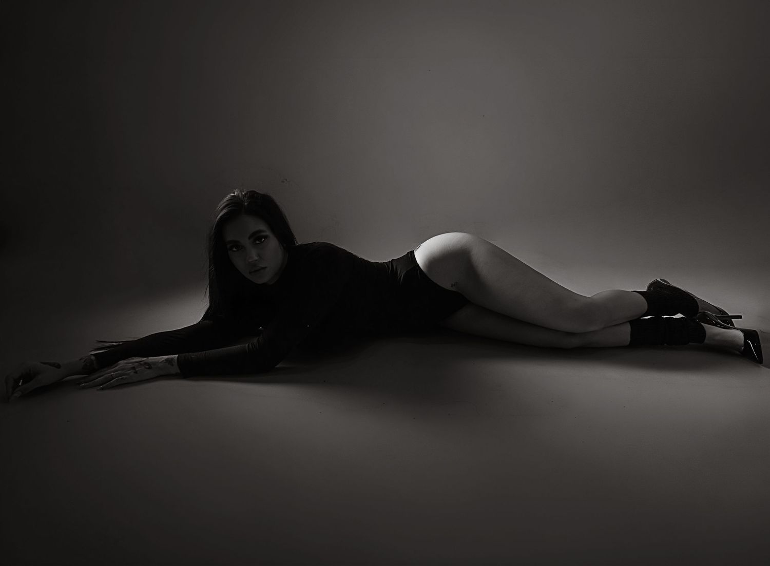 Black and white studio photo of a woman in black bodysuit lying gracefully on the floor in a dramatic pose.