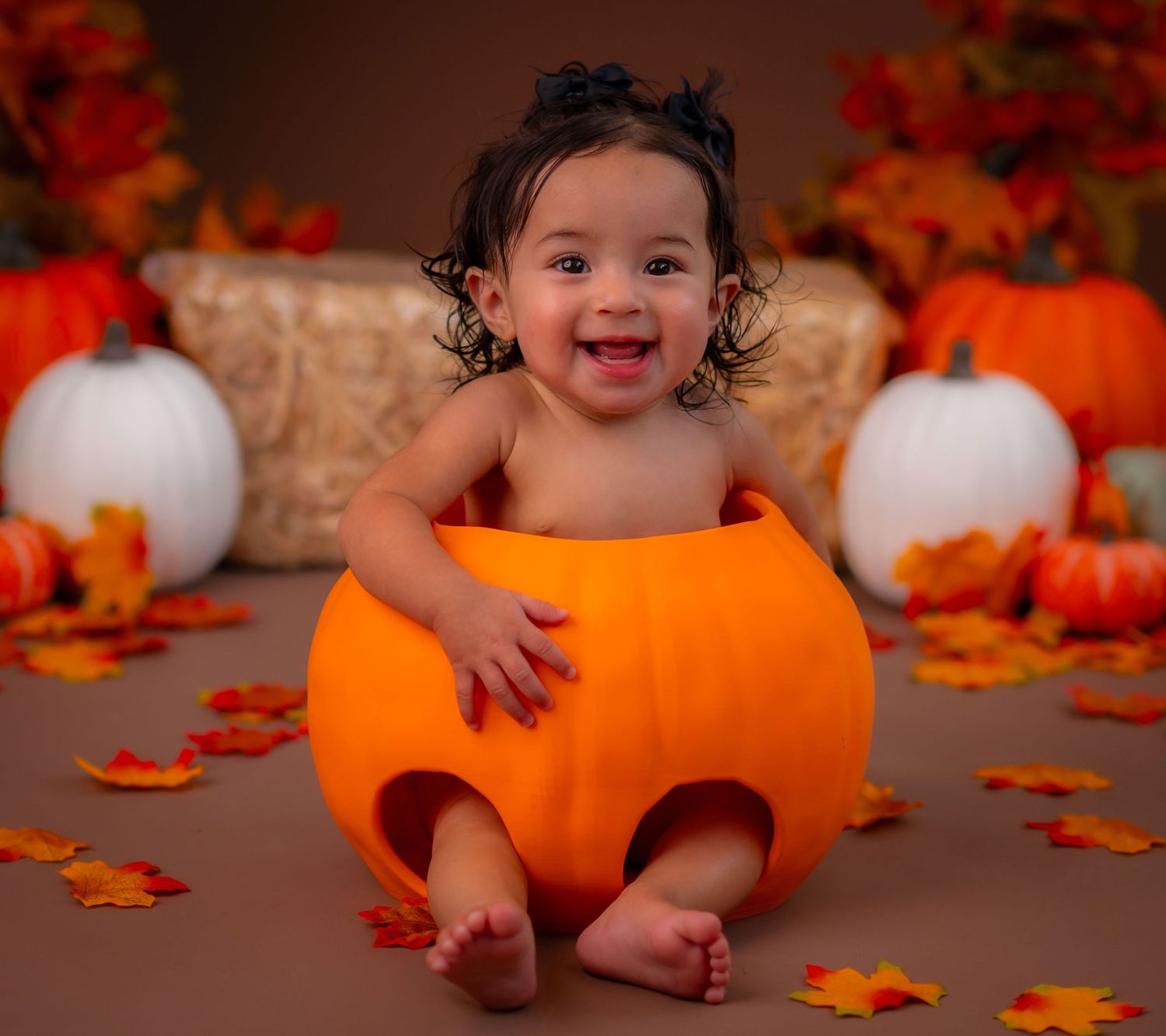 Smiling baby sitting inside a large orange pumpkin surrounded by fall leaves and pumpkins in a Halloween photo shoot.