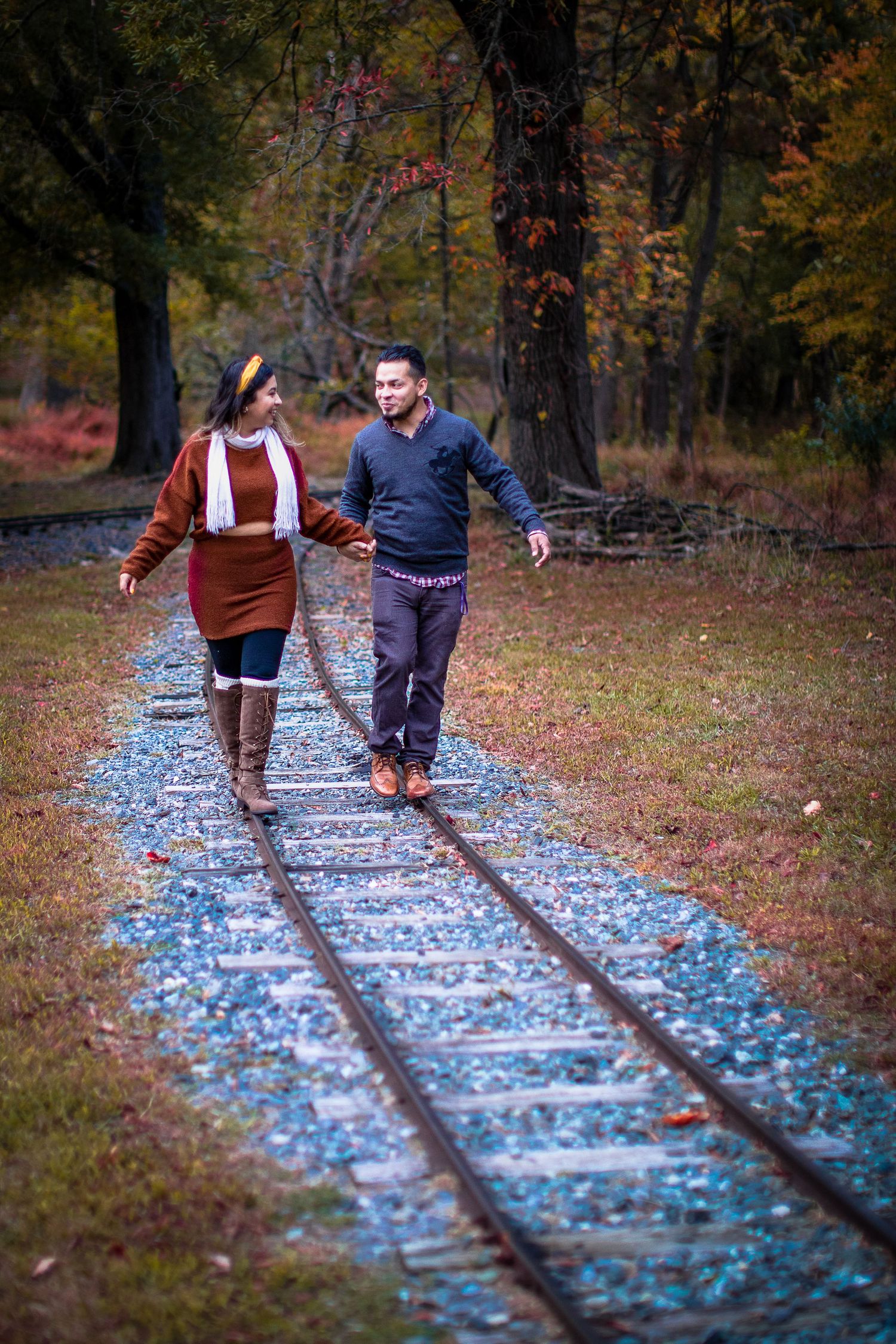 Couple walking hand-in-hand on railroad tracks surrounded by colorful autumn foliage in a wooded setting.