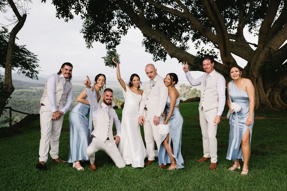Wedding party posing joyfully under a large tree in blue bridesmaid dresses and white suits.