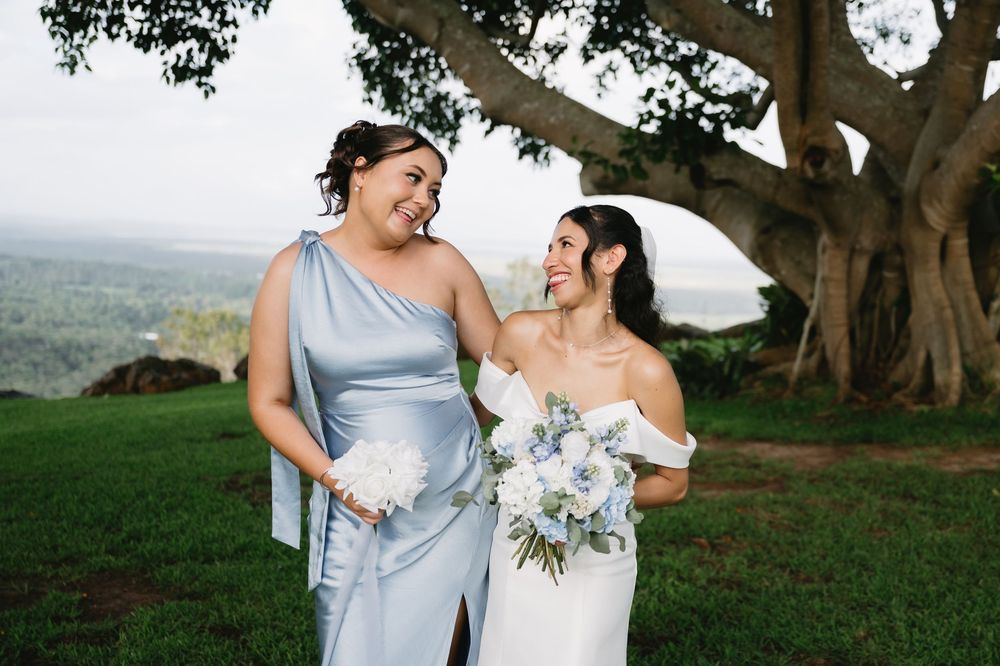 Bride in white off-shoulder gown and bridesmaid in blue dress smiling together outdoors near large tree.