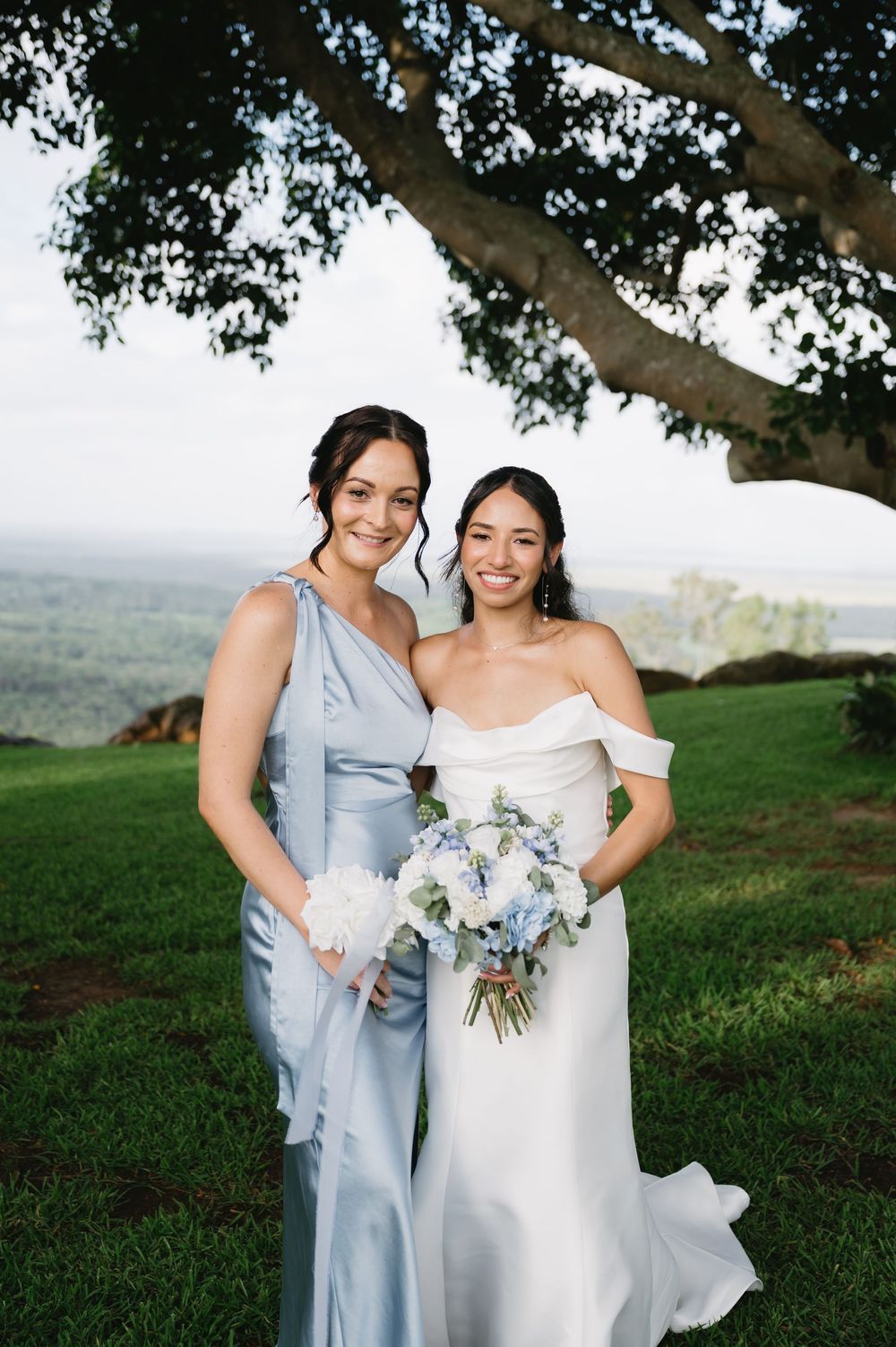 Bride in white off-shoulder gown holds blue bouquet beside bridesmaid in dusty blue dress, outdoors under a tree.