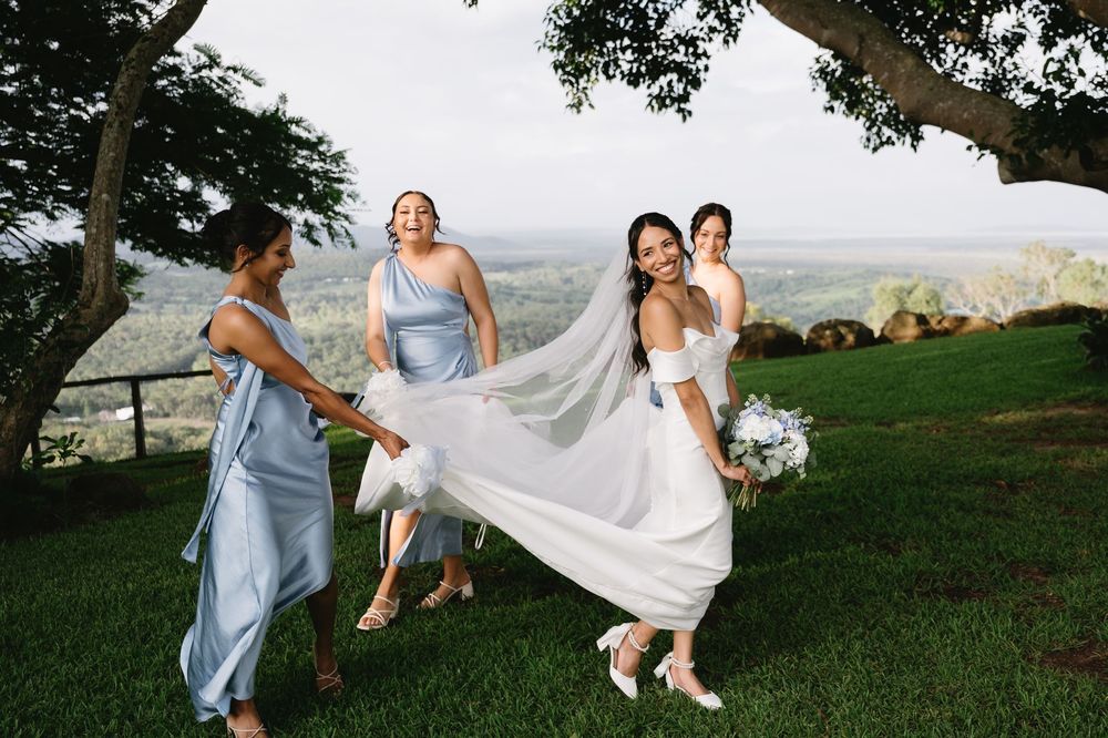 Bride in white gown with veil walks with bridesmaids in blue dresses on green hillside with scenic valley views.