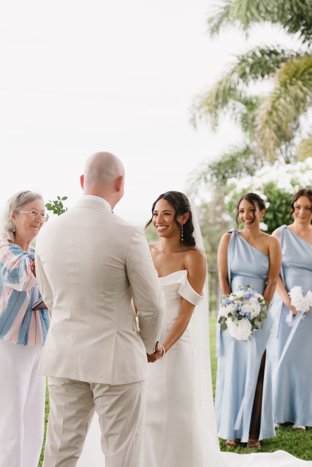 Bride and groom hold hands at outdoor wedding ceremony with bridesmaids in blue dresses and officiant nearby.
