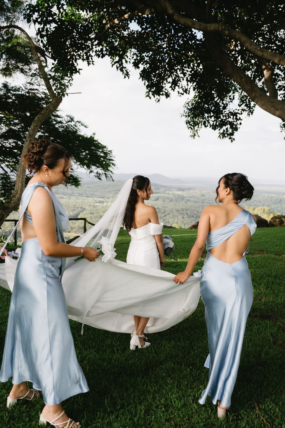 Two bridesmaids in blue satin dresses hold a bride's veil outdoors at a scenic hilltop wedding ceremony.