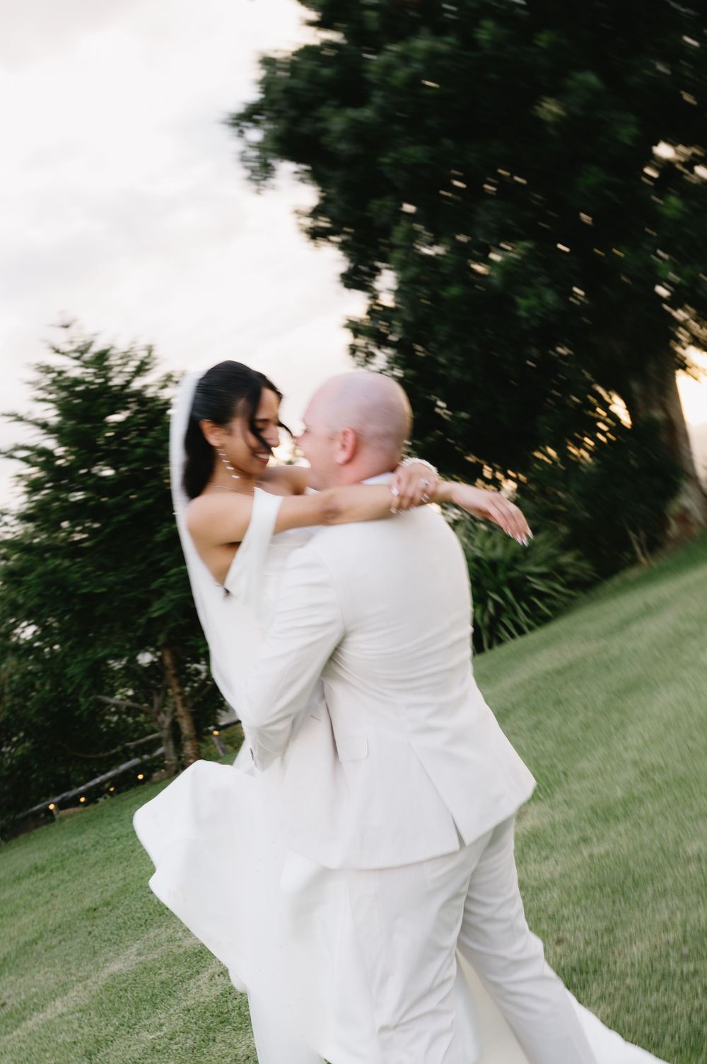 A groom in a white suit lifts his bride in a flowing white gown outdoors on a green lawn at sunset.