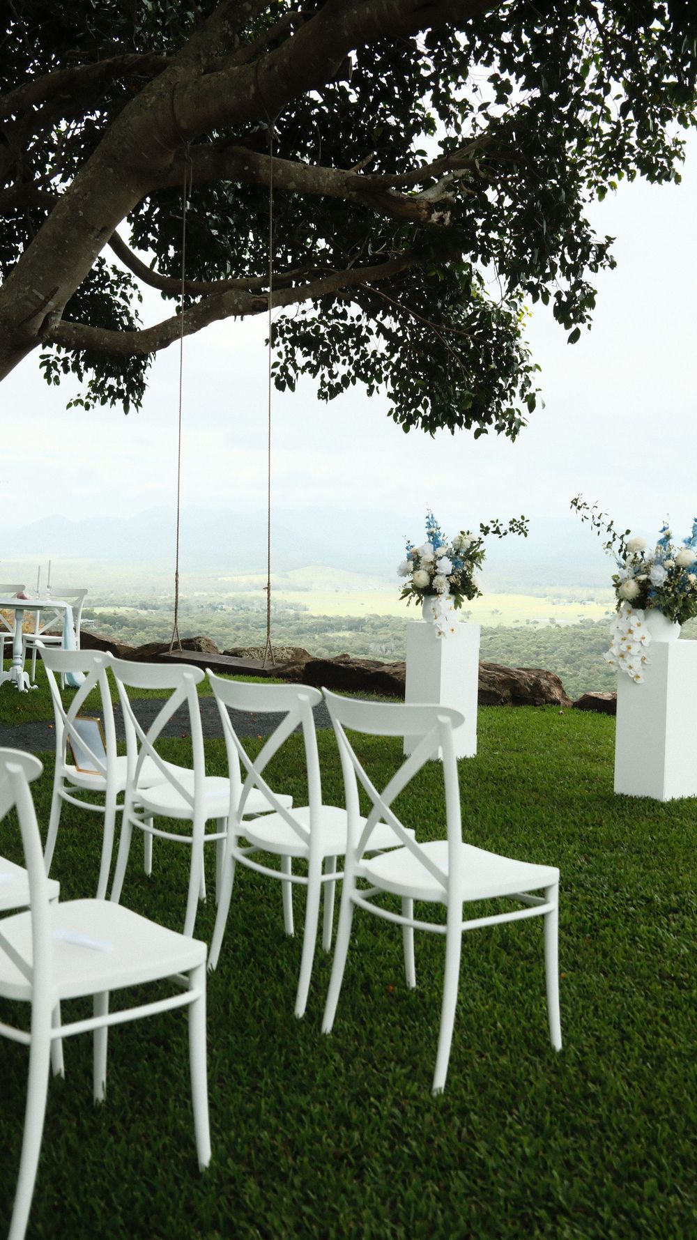 White cross-back chairs arranged on green grass under a tree for an outdoor wedding ceremony with floral arrangements and scenic views.