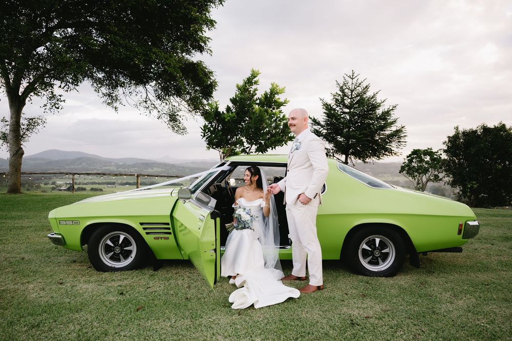 Bride and groom posing beside a classic green muscle car on lush grass with scenic mountain views.