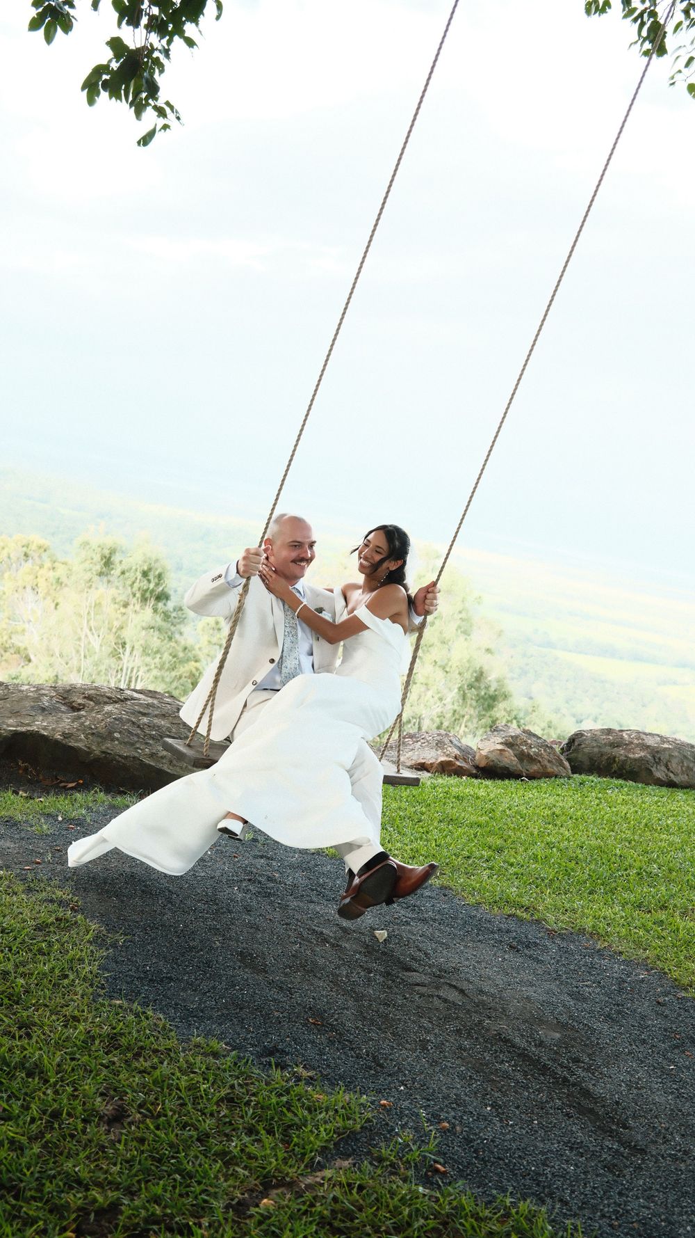Bride and groom in white attire laughing together on a large rope swing with scenic green hills in the background.