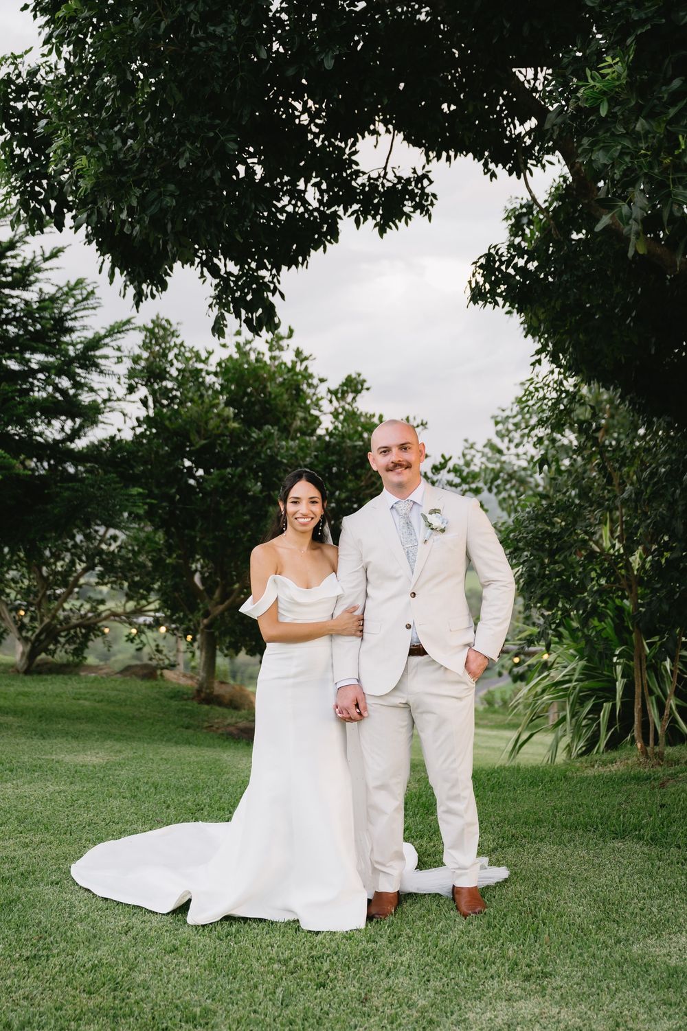 Bride in off-shoulder white gown and groom in beige suit pose together on lush green lawn under leafy trees.