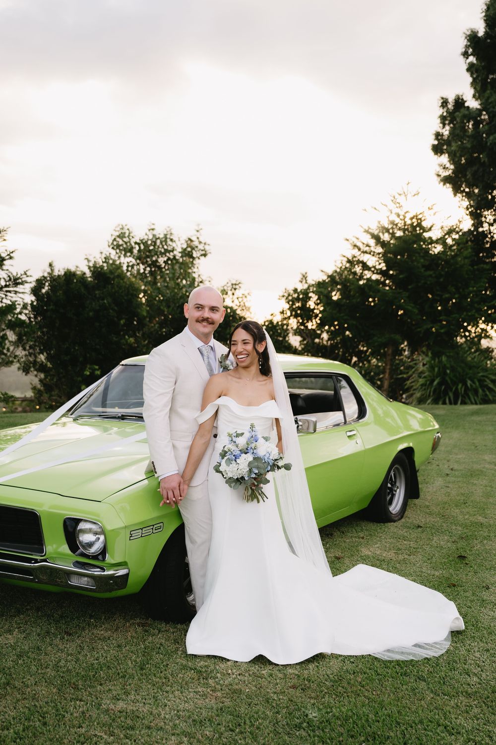 Bride and groom posing in front of a bright green classic car on their wedding day outdoors.
