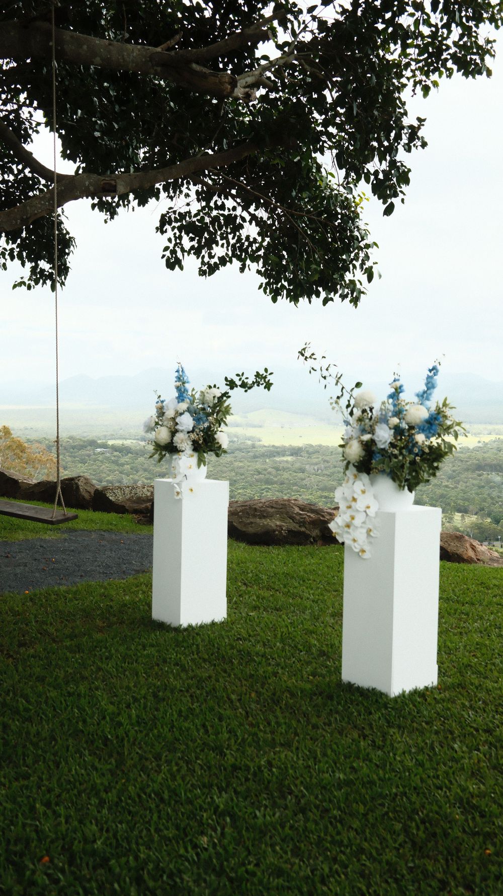 Two white pedestal stands with blue and white floral arrangements on green lawn overlooking scenic valley landscape.