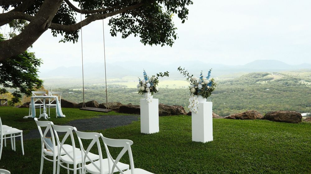 Outdoor wedding ceremony setup with white chairs, floral arch, and stunning valley views under a large tree.