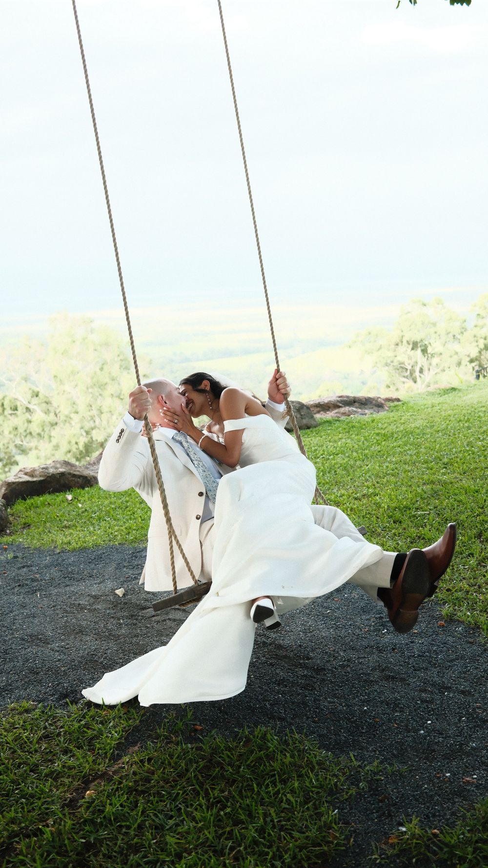 Bride and groom kissing on a rope swing outdoors, dressed in white wedding attire with scenic green landscape behind them.